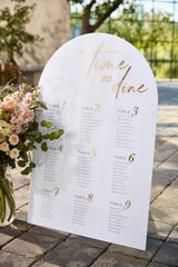 White acrylic arched seating chart with gold mirror text displayed at wedding reception
