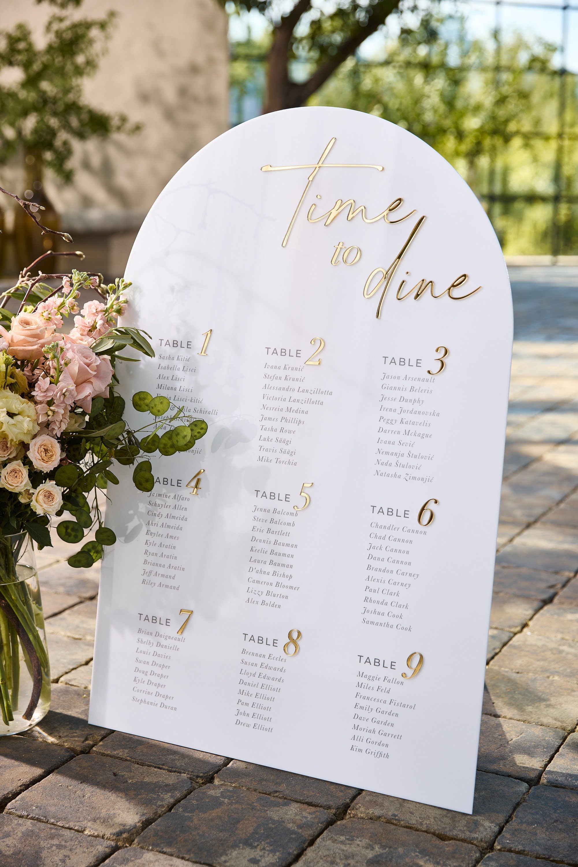 White acrylic arched seating chart with gold mirror text displayed at wedding reception
