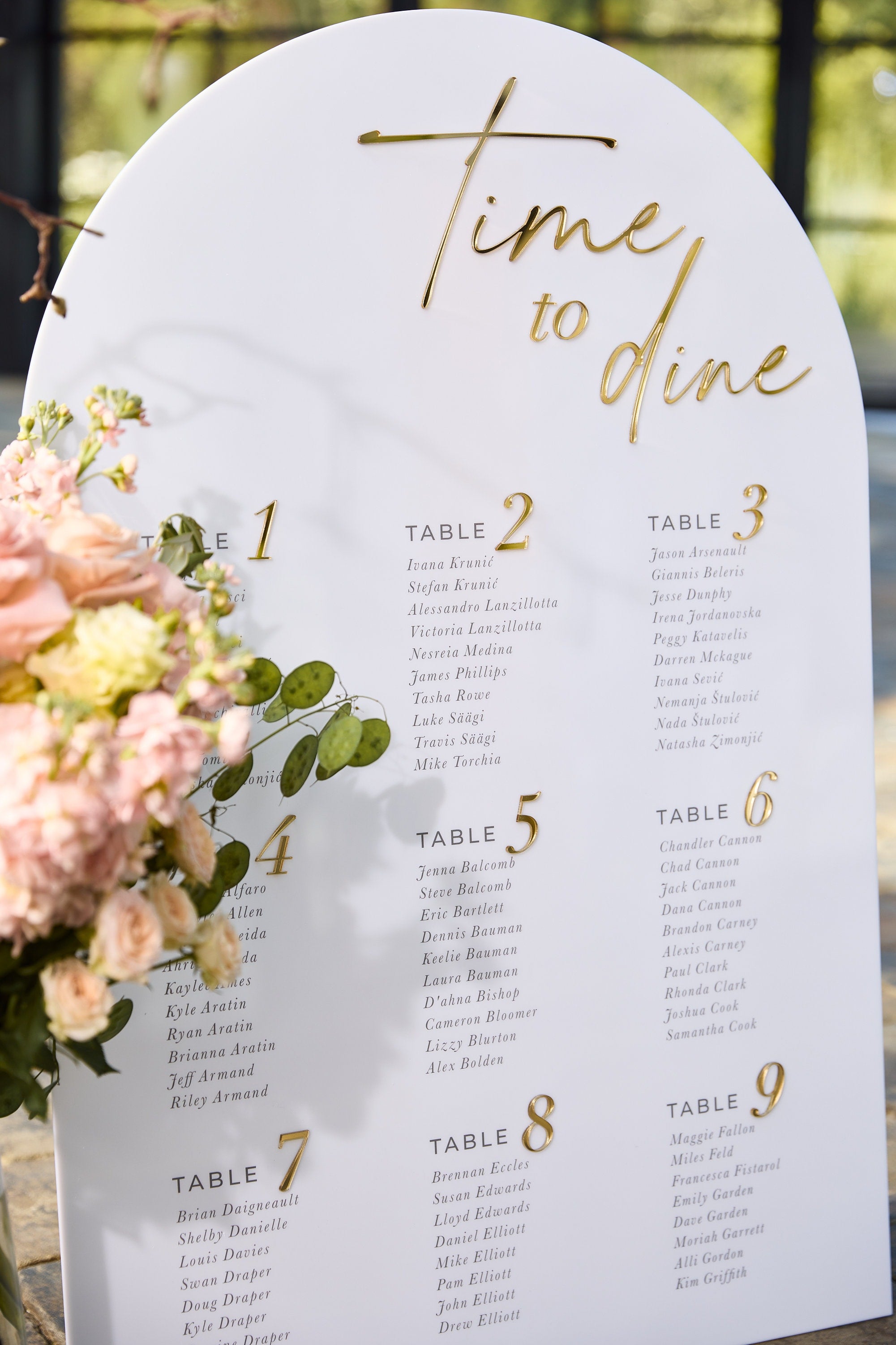 Arched acrylic seating chart with gold mirror lettering displaying multiple table assignments
