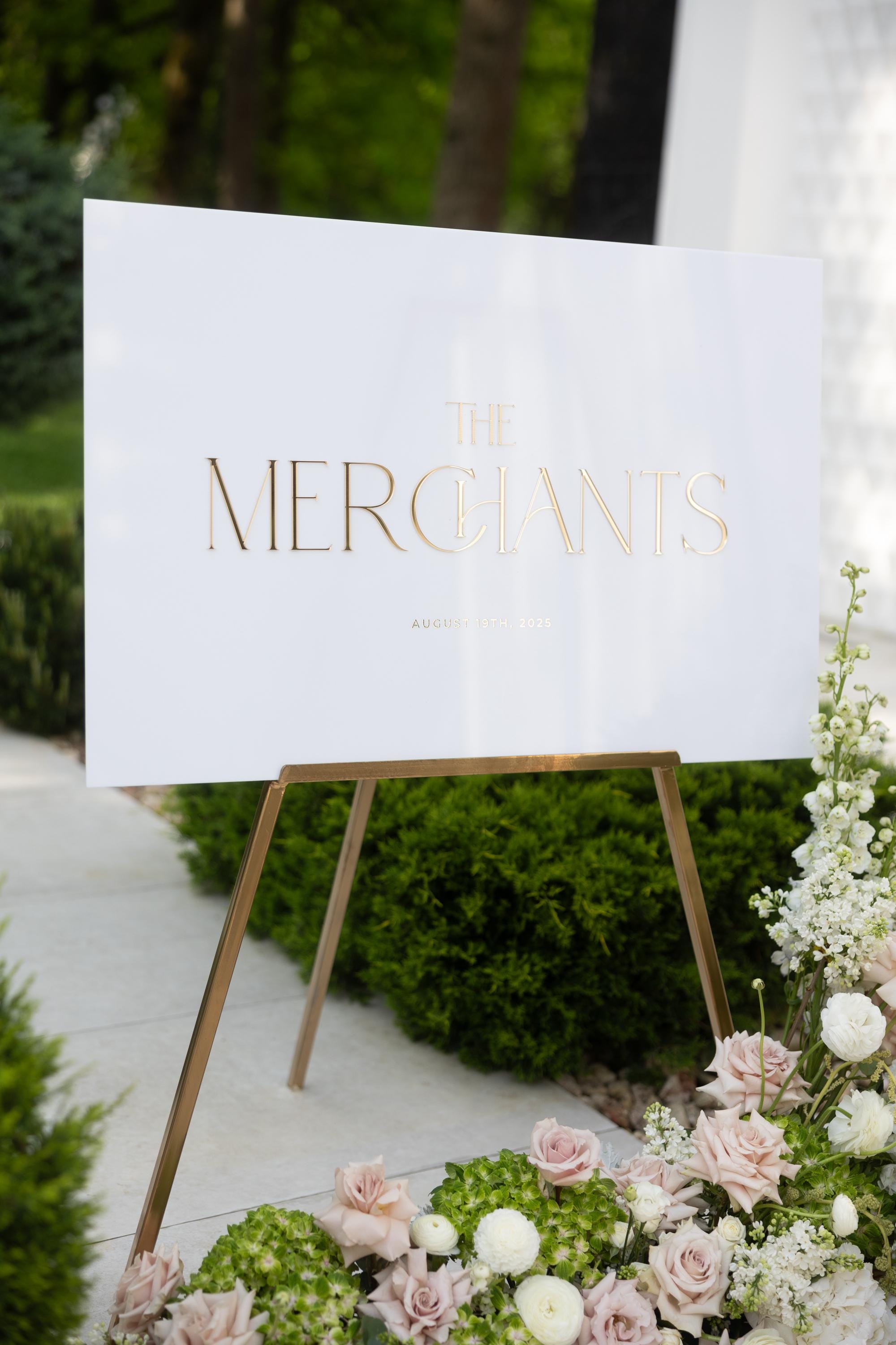Elegant white and gold wedding signage displayed on an easel at ceremony entrance.